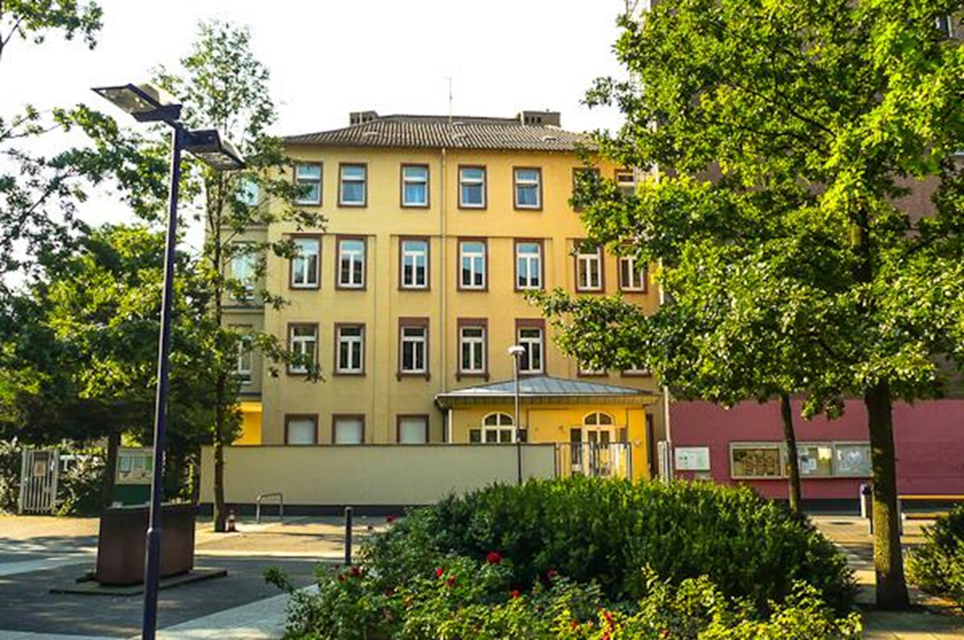 Auf dem Bild sieht man die Außenansicht des Caritashauses St. Josef.
Es handelt sich um ein mehrstöckiges Gebäude mit heller, gelblicher Fassade.
Das Haus hat mehrere Reihen von Fenstern, alle gleichmäßig angeordnet.
Links und rechts stehen hohe Bäume, die teilweise das Gebäude verdecken.
Vor dem Haus befindet sich ein kleiner Platz mit Sträuchern, Blumen und Straßenlampen.