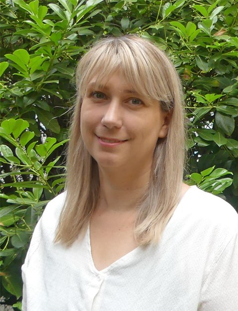 Porträtfoto einer jungen Frau mit halblangem blonden Haar, die eine kurzärmelige helle Bluse trägt. Im Hintergrund ist grünes Blattwerk zu sehen.